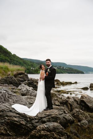 Bride and groom embracing on rocky shoreline with mountain lake backdrop on their wedding day.