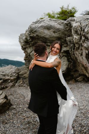 Smiling bride in white gown embracing groom near rocky coastal cliffs on their wedding day.