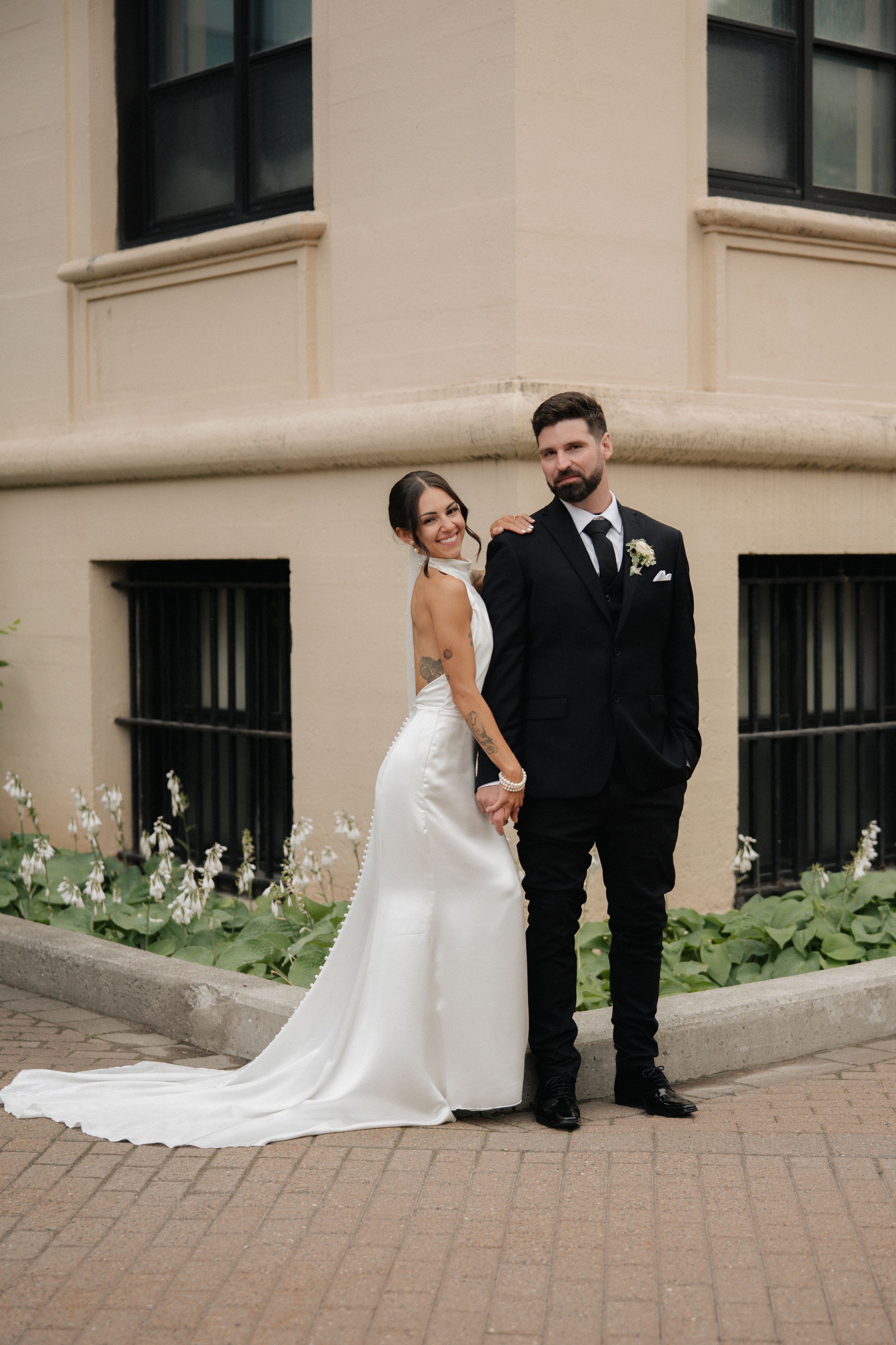 Bride in white halter gown and groom in black tuxedo hold hands outside a beige building on their wedding day.