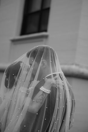 Black and white photo of couple kissing under pearl-adorned bridal veil outdoors near building.