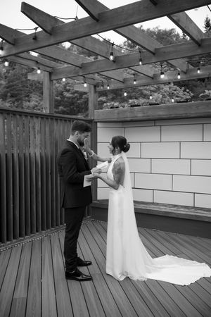 Black and white photo of bride and groom exchanging vows on a wooden deck pergola with string lights.