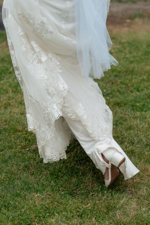 Close-up view of white wedding dress hem and elegant bridal shoes walking on green grass.