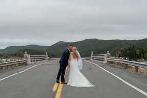 A couple shares a romantic moment on a mountain road bridge with scenic forested hills in the background.