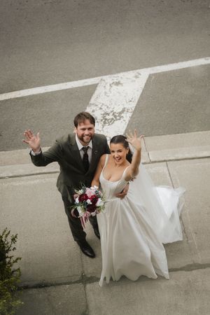 Bride and groom waving from above on wedding day, bride holding bouquet in white gown, groom in gray suit.