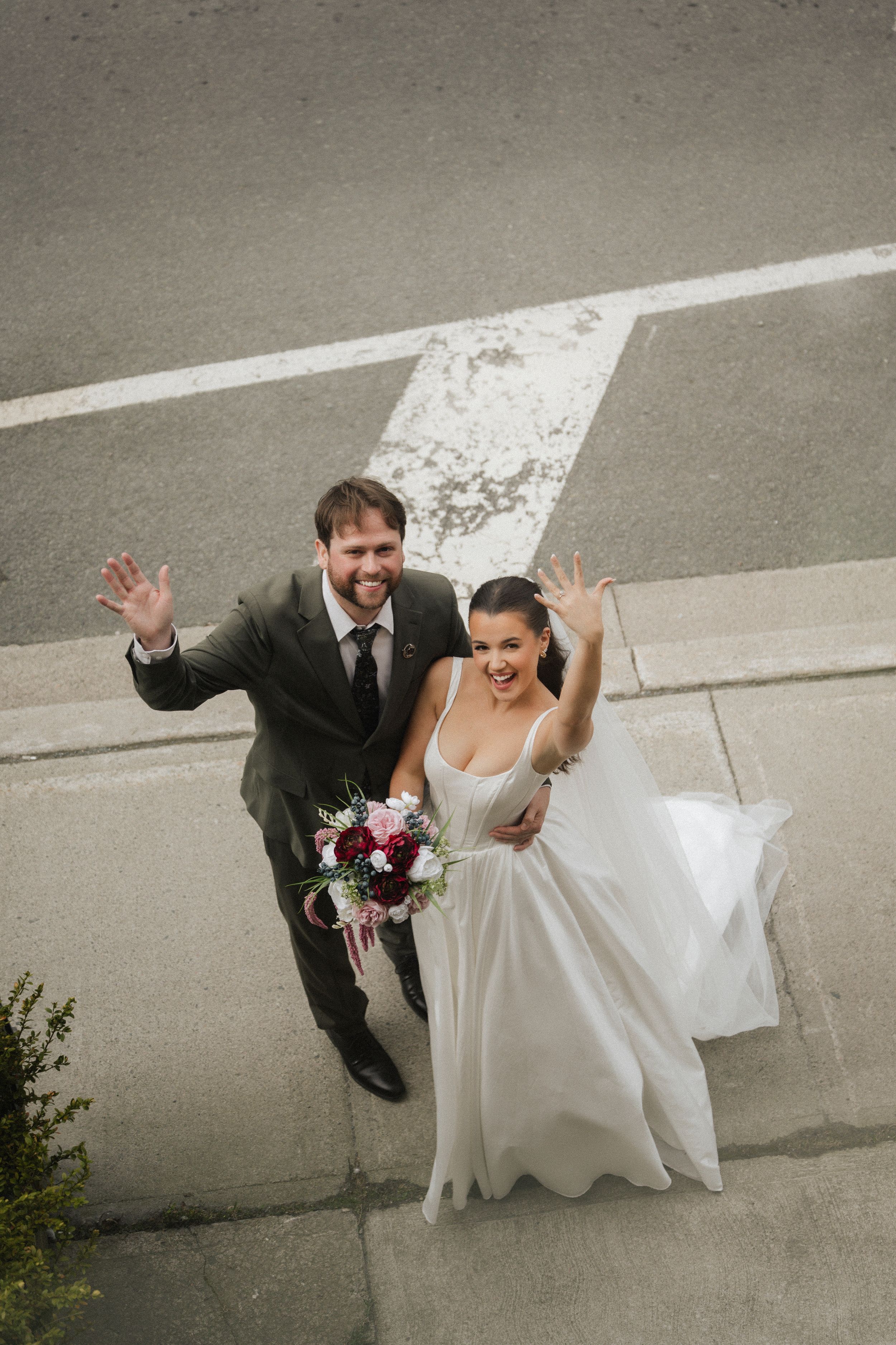 Bride and groom waving from above on wedding day, bride holding bouquet in white gown, groom in gray suit.