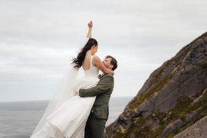 Bride and groom share romantic dance on rocky coastal cliff, bride in white gown with arm raised, groom in olive suit.