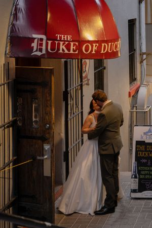A bride and groom share a romantic kiss under the red Duke of Duck awning in an intimate urban setting.