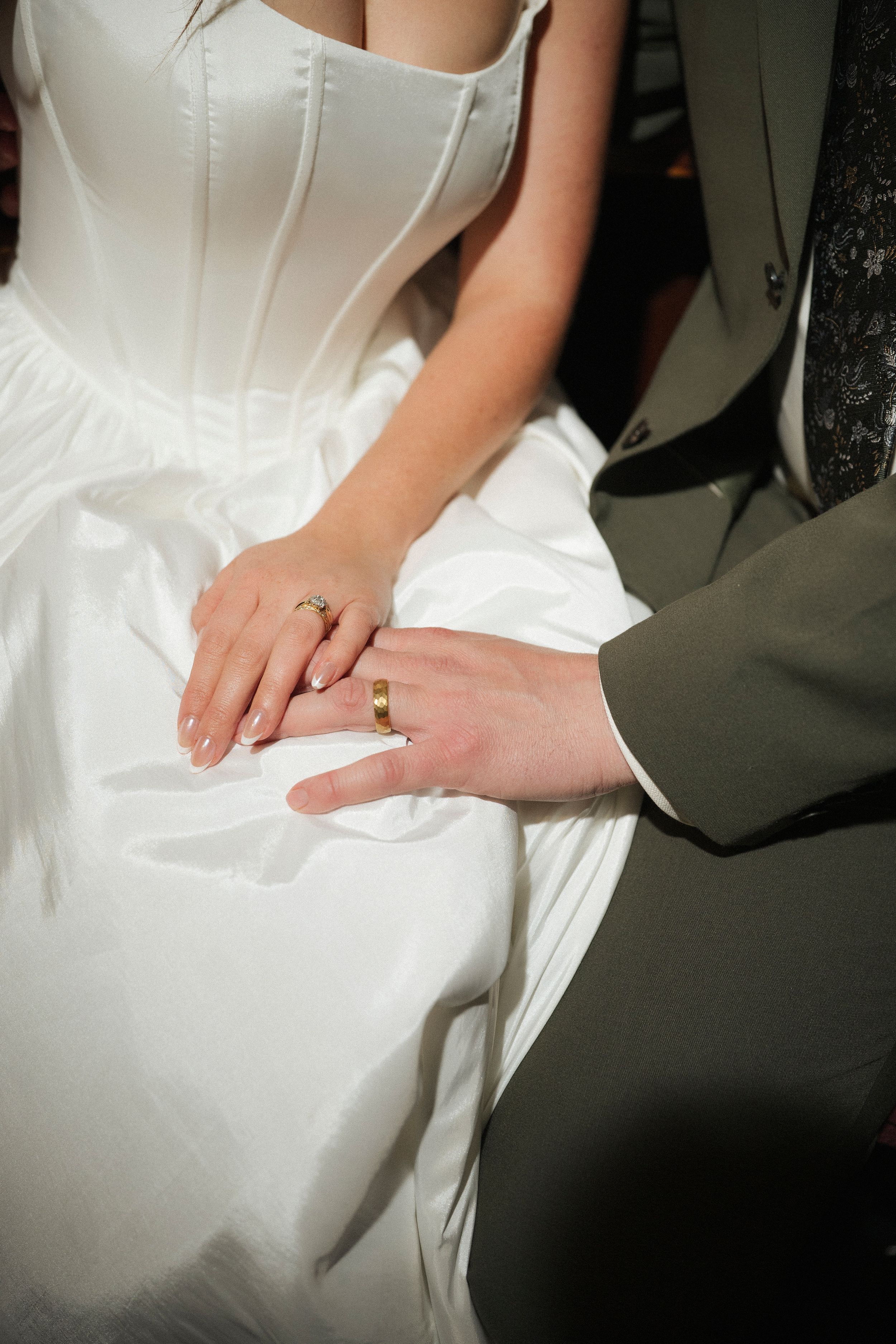 Bride and groom holding hands showing gold wedding rings, bride in white gown, groom in gray suit.