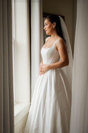 Elegant bride in white corset ballgown and veil gazing out window on wedding day.