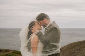 Bride and groom sharing a romantic moment under a flowing veil with ocean cliffs in the background.