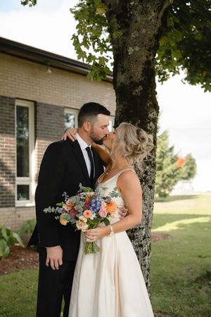 A couple shares a romantic moment under a tree, with the bride holding a colorful bouquet of flowers.