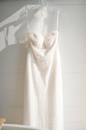 White lace spaghetti strap wedding dress with sweetheart neckline hanging on clear hanger against white shiplap wall.