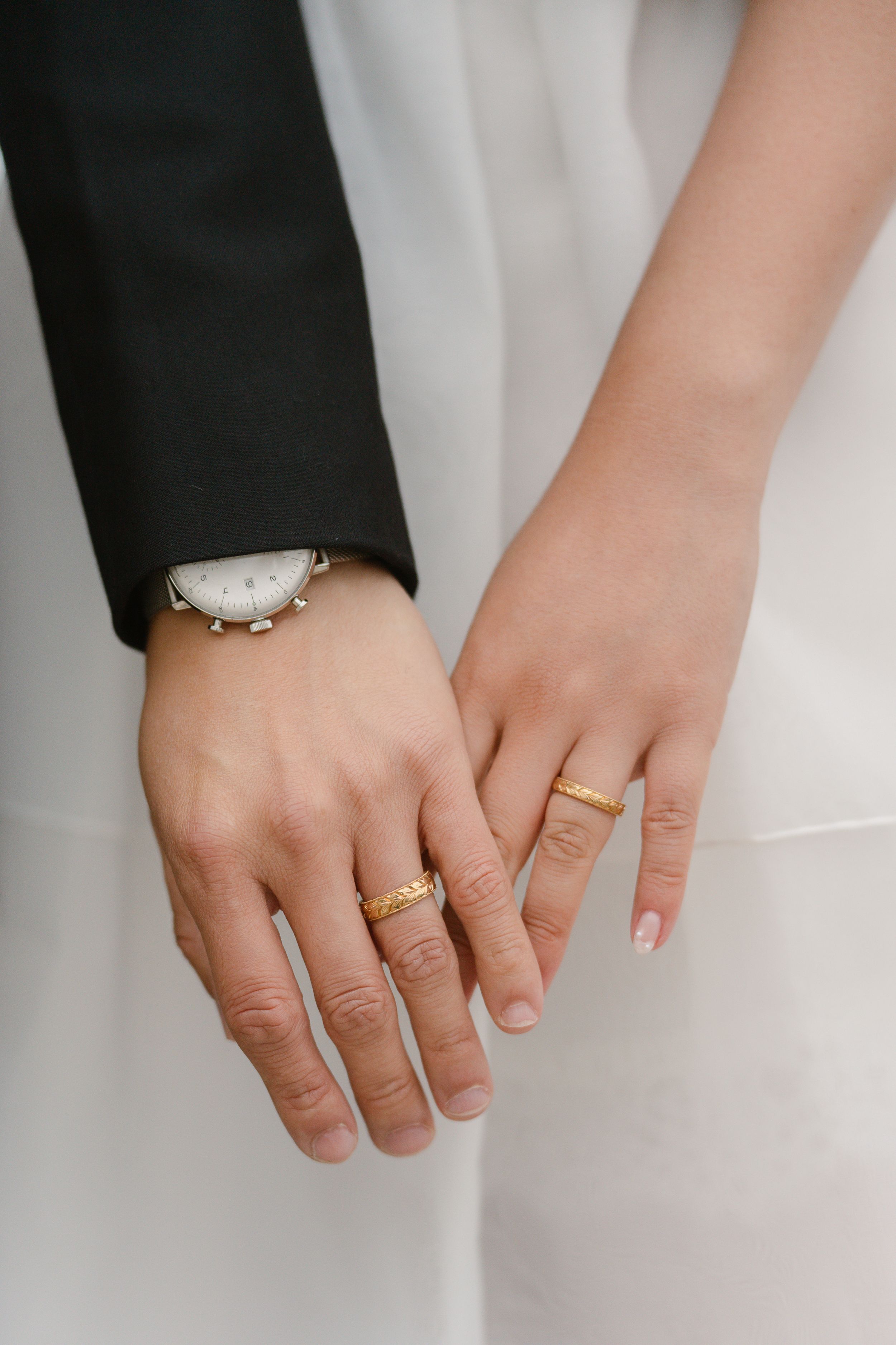 Bride and groom holding hands displaying matching gold wedding bands, with groom wearing a white watch and black suit.