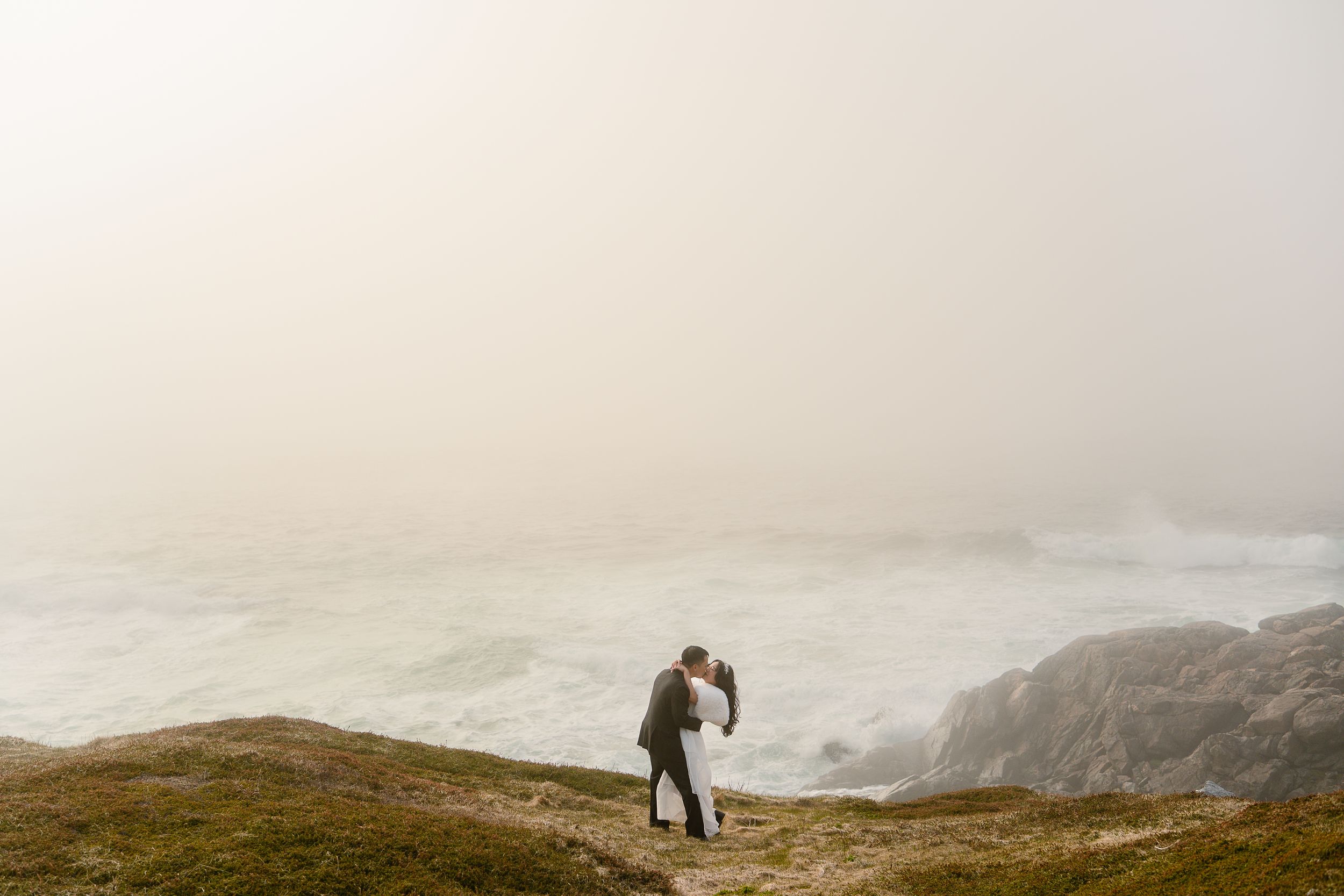 Couple sharing a romantic kiss on a misty coastal cliff with dramatic ocean waves crashing below.