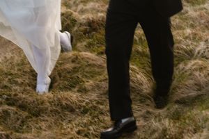 Bride and groom walking through grassy field, white wedding dress and black suit visible from waist down.