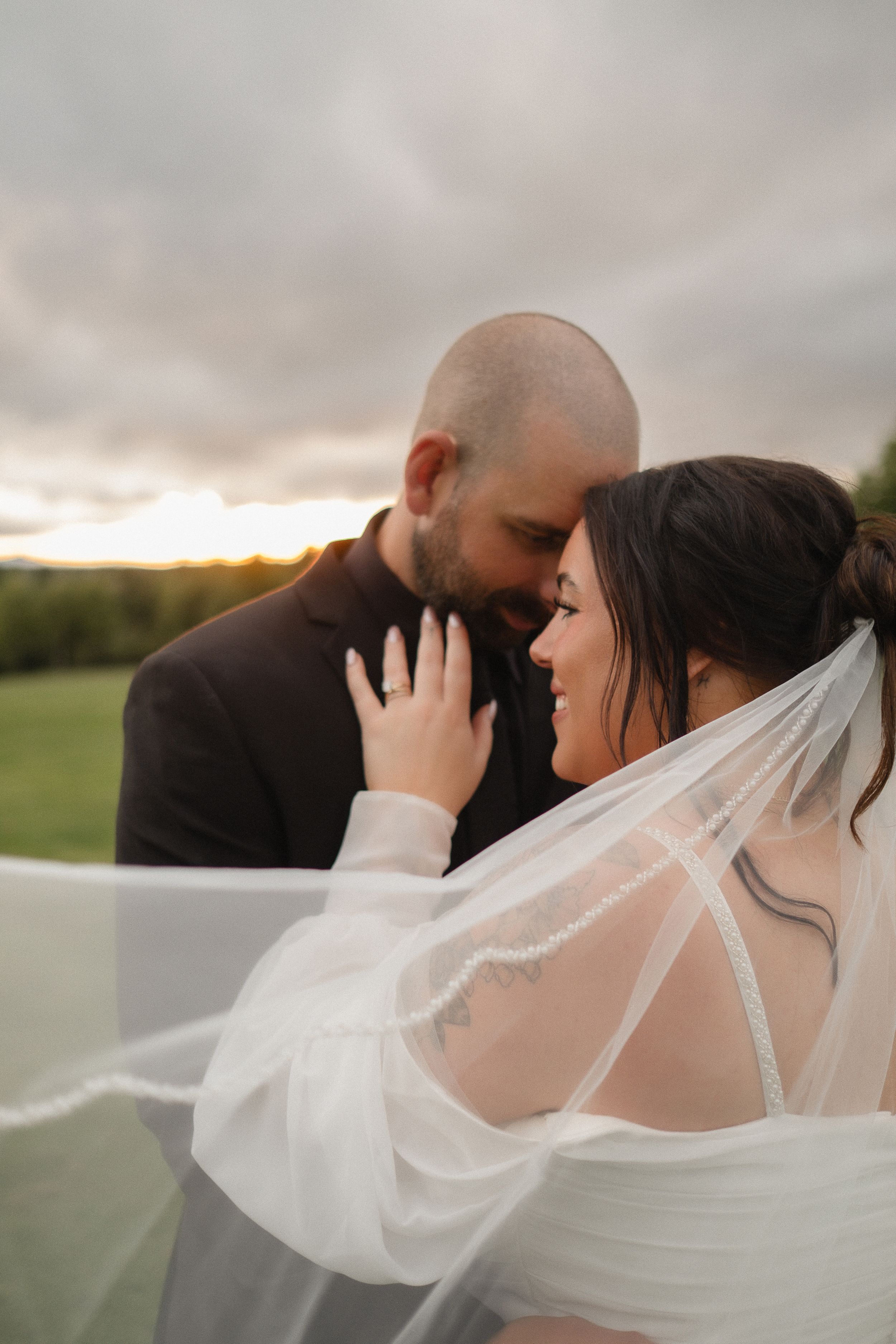A romantic wedding couple embraces in a field at sunset as the bride's veil flows dramatically in the wind.