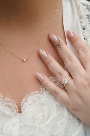 A delicate gold necklace with a small pearl pendant rests against white lace fabric, paired with manicured hands and rings.
