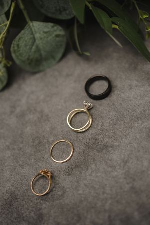 A series of golden engagement rings with diamonds photographed on a grey stone surface with green foliage accents.