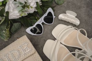 Wedding accessories including heart-shaped sunglasses, hair clip, bridal shoes and beaded clutch arranged with white flowers.