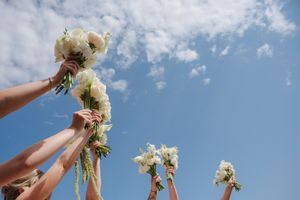 Bridesmaids raising white flower bouquets against a blue sky at a wedding celebration outdoors.
