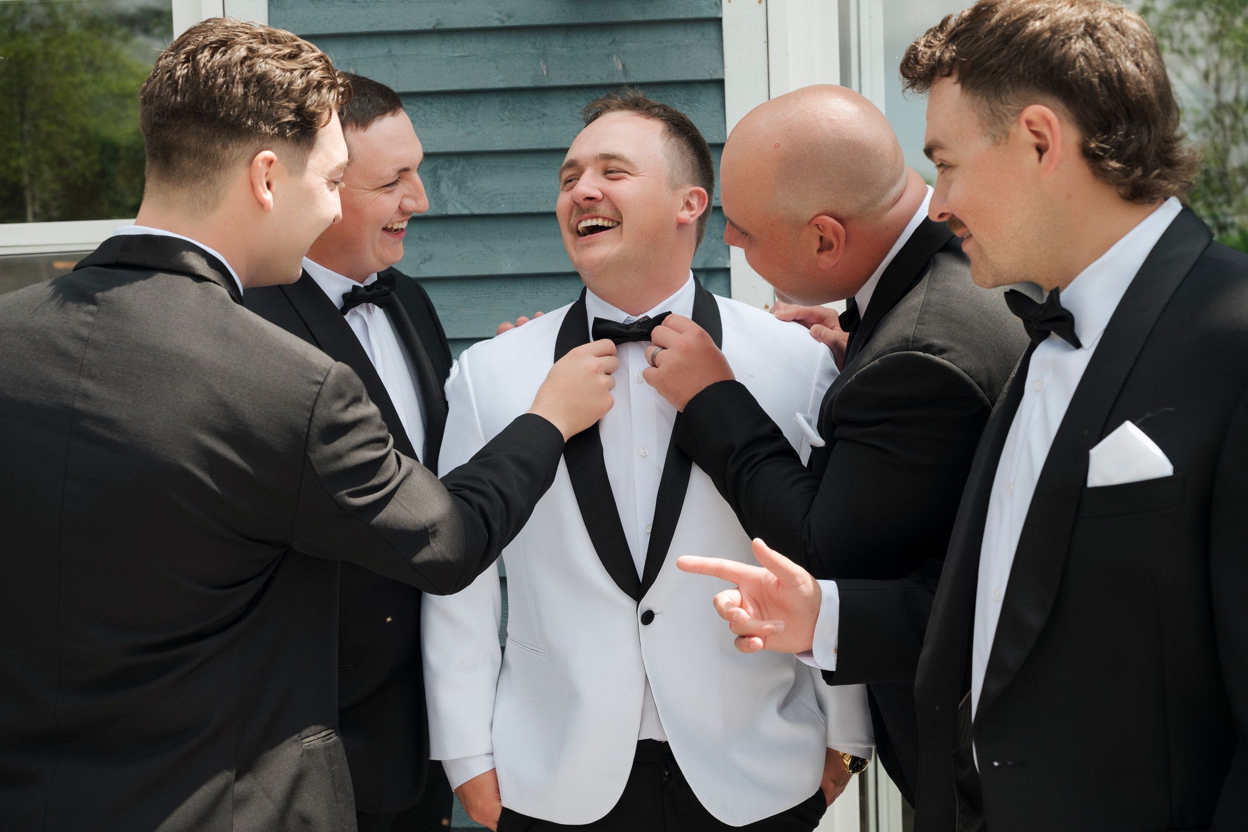 Groom in white tuxedo jacket laughs with groomsmen in black suits adjusting his bow tie before wedding ceremony.