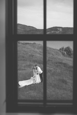 Black and white photo of bride and groom embracing on a green hillside, viewed through a window frame.