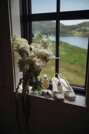 Bridal bouquet of white roses and calla lilies with heels, perfume, and accessories on a window sill overlooking a scenic lake.