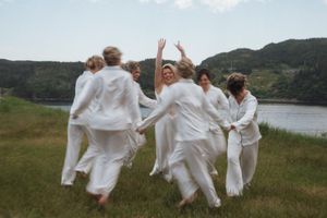Group of women in white dresses dancing joyfully on green grass near a scenic lake and hills.