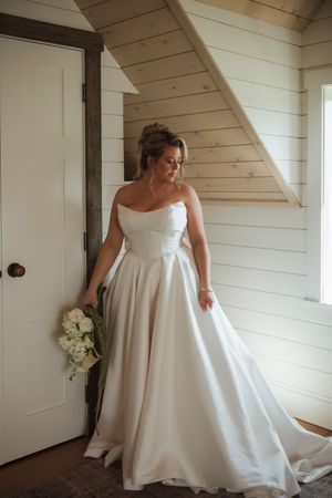 Bride in strapless white satin ballgown holding white bouquet, standing in rustic wood-paneled room with natural light.