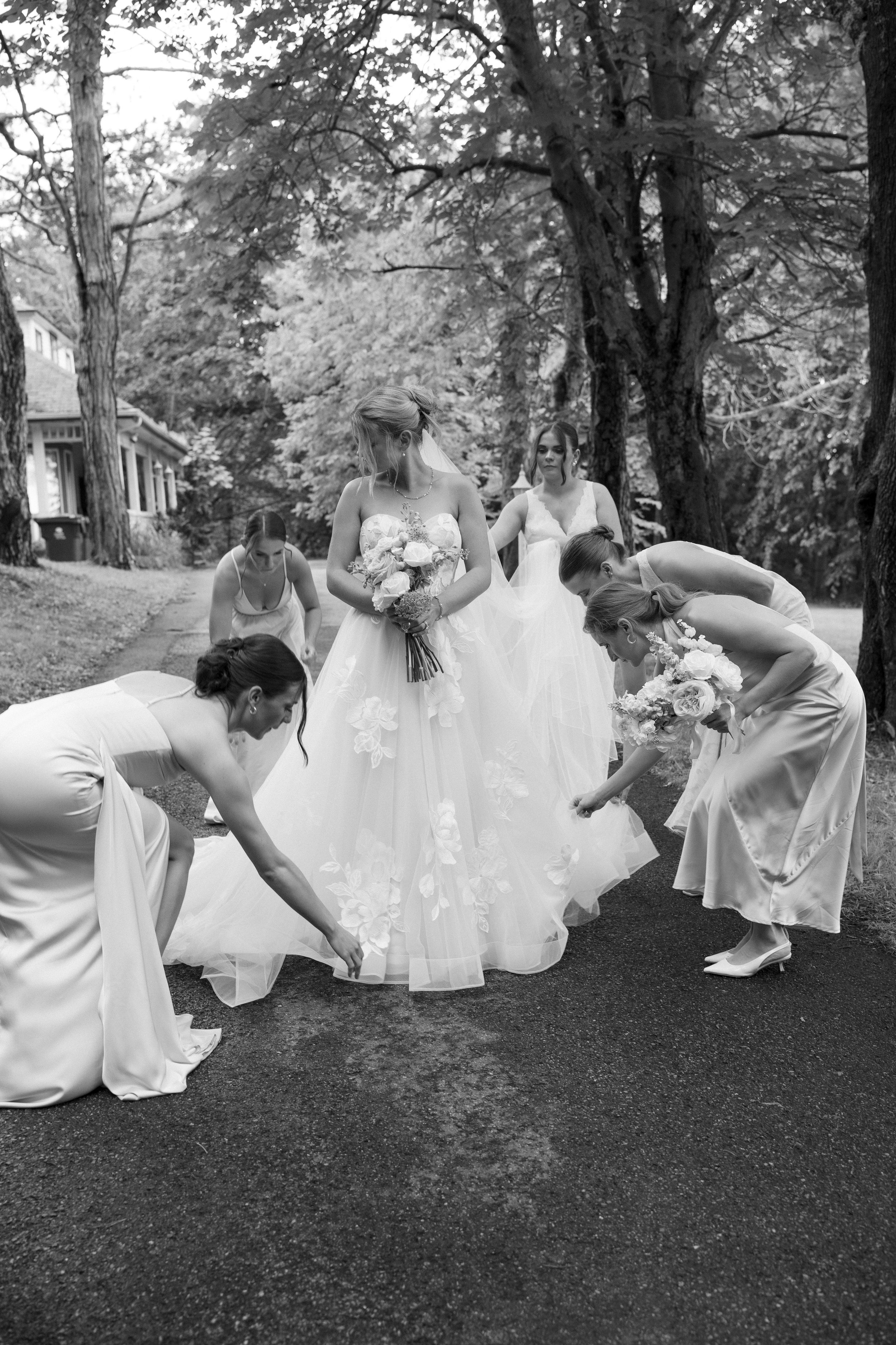 Bridesmaids in white gowns adjust the bride's floral wedding dress outdoors, surrounded by trees, in a black and white photo.