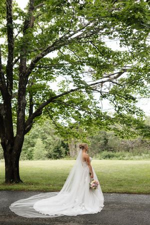 Bride in a long white gown and veil stands beneath a large green tree, holding a bouquet on a lush lawn.
