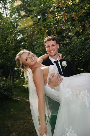Groom in black suit carries smiling bride in white lace wedding dress outdoors in lush green garden.