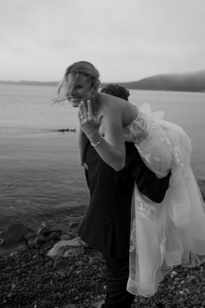 Black and white photo of a groom carrying his laughing bride by the rocky shoreline on their wedding day.