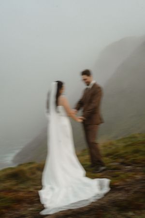 A couple in wedding attire stands on a misty cliffside overlooking the ocean in a dreamy, atmospheric scene.