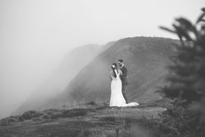 A couple in wedding attire stands on a misty mountain ridge overlooking a dramatic coastal landscape at sunset.
