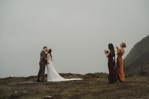 Wedding couple stands on misty coastal cliff while photographers capture their intimate moment in moody lighting.