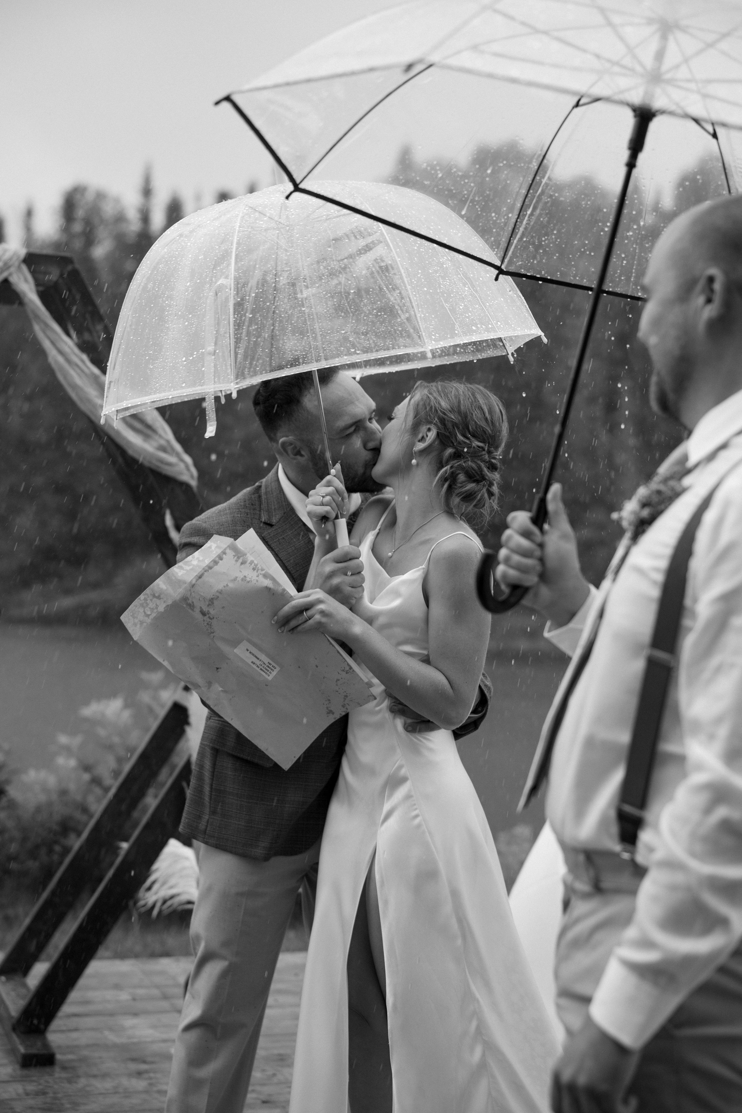 Black and white photo of a bride and groom kissing in the rain under a clear umbrella on their wedding day.