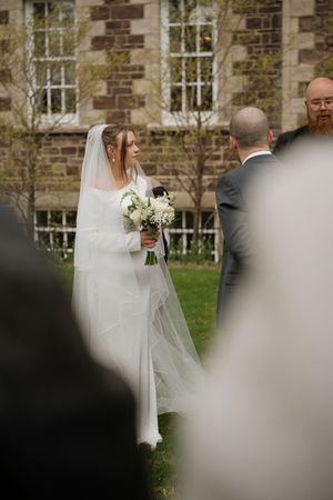 Bride in white gown and veil holds bouquet during outdoor wedding ceremony near stone building.
