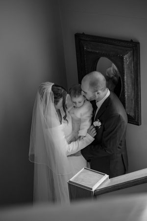 Black and white wedding photo of bride and groom holding baby near mirror and registry book.