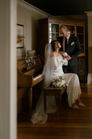 Bride in white gown and veil sits at piano while groom in dark suit holds her hand in elegant home interior.