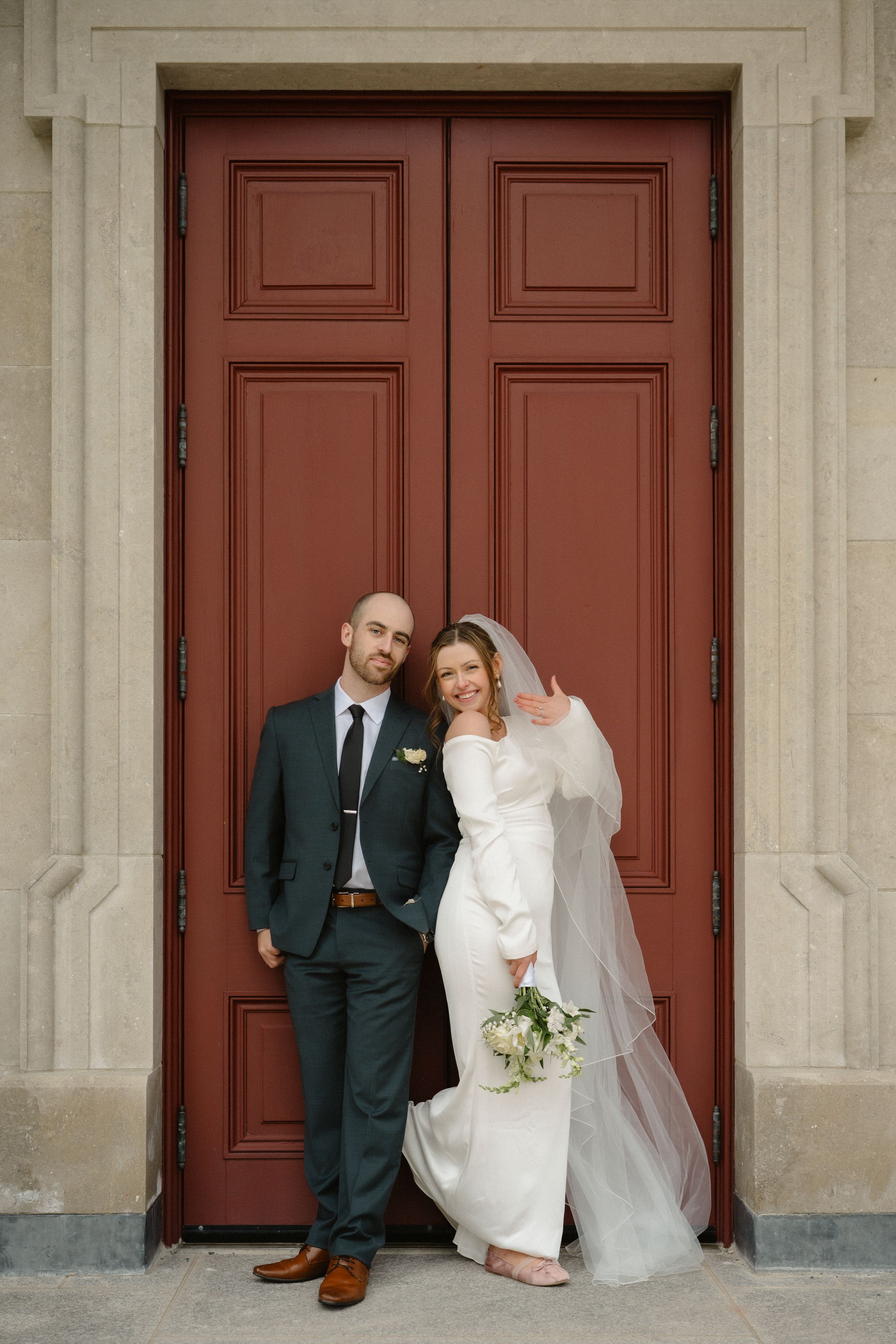 Bride and groom posing in front of large red doors, bride holding white bouquet and wearing veil, groom in dark suit.