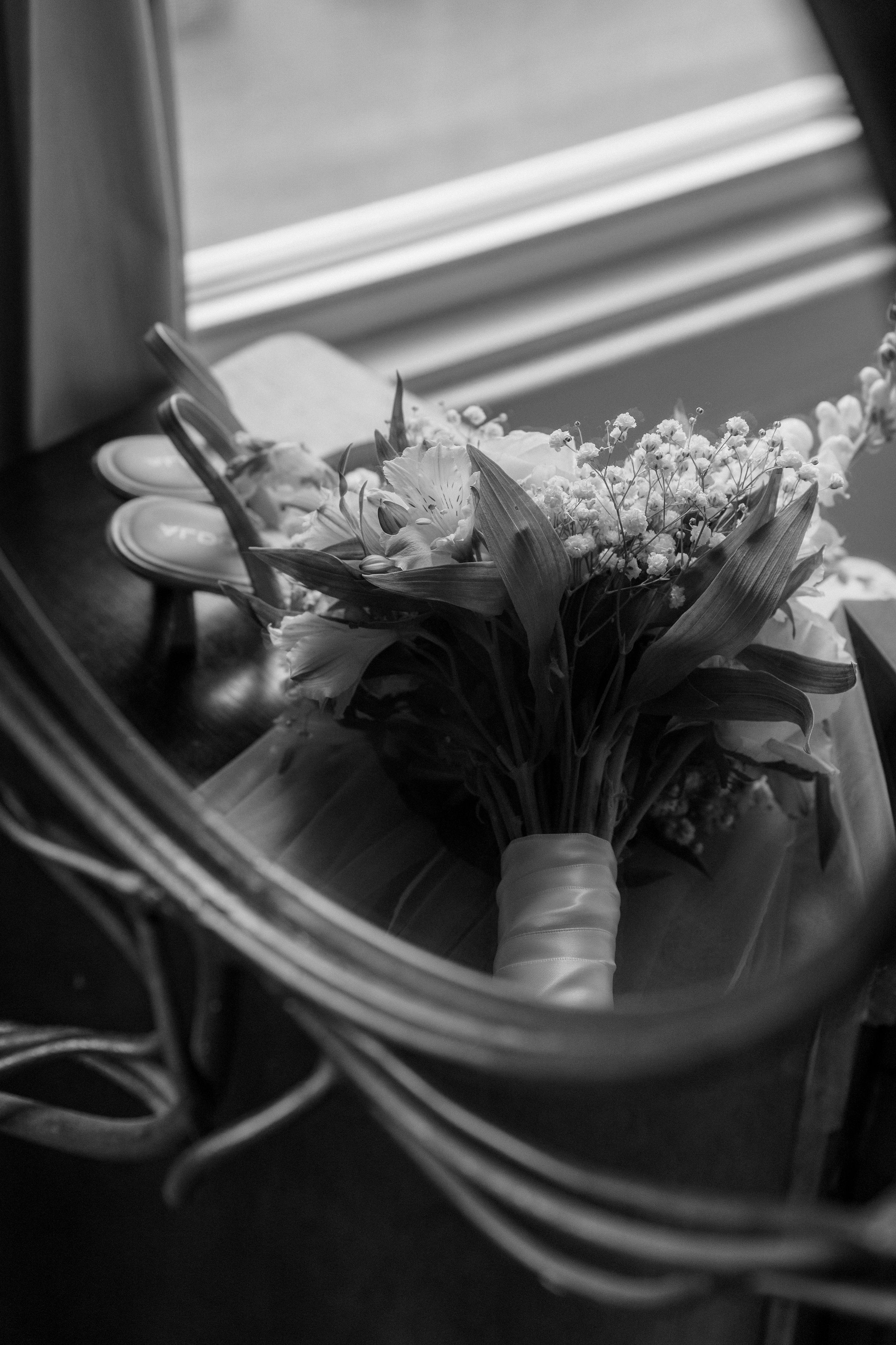 Black and white photo of a bridal bouquet with ribbon resting on a chair near a sunlit window.