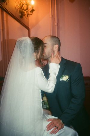 Bride and groom share a romantic kiss at their wedding, with the bride in a white gown and veil, groom in a navy suit.