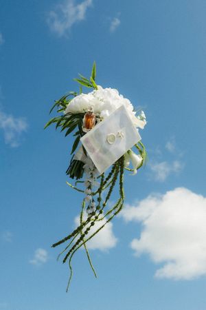 Wedding bouquet of white flowers tossed against a bright blue sky with fluffy clouds.