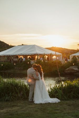 Bride and groom share a romantic sunset kiss by a pond with a white wedding tent glowing in the golden hour light.