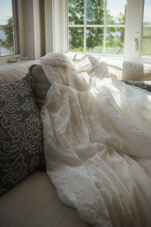 White lace wedding dress draped elegantly over a couch with decorative pillows near a bright window.