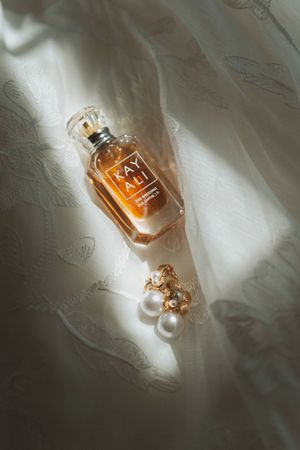 Kayali perfume bottle with gold pearl earrings on white lace fabric in soft natural light.