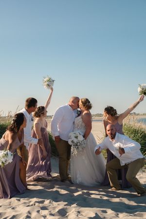 A wedding party celebrates joyfully on a sandy beach with bridesmaids in purple dresses holding bouquets against a blue sky.