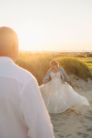 A couple sits together on sandy beach dunes during a romantic golden hour sunset photoshoot.