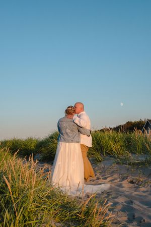 A couple sits together on a sandy beach path surrounded by dune grass at sunset with a soft blue sky.